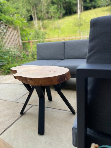 Wooden side table with a natural edge, surrounded by outdoor furniture in a garden setting.