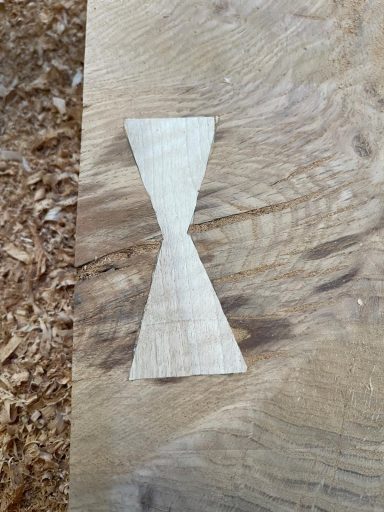 Wooden piece shaped like an hourglass, placed on a wood surface with shavings.