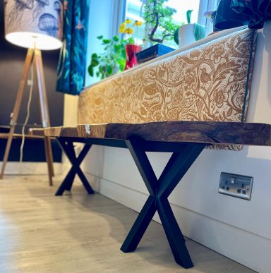 Stylish wooden bench with black X-shaped legs, set in a bright room with plants.