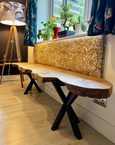 Wooden bench with a natural edge, supported by black metal legs, beside a lamp and plants.